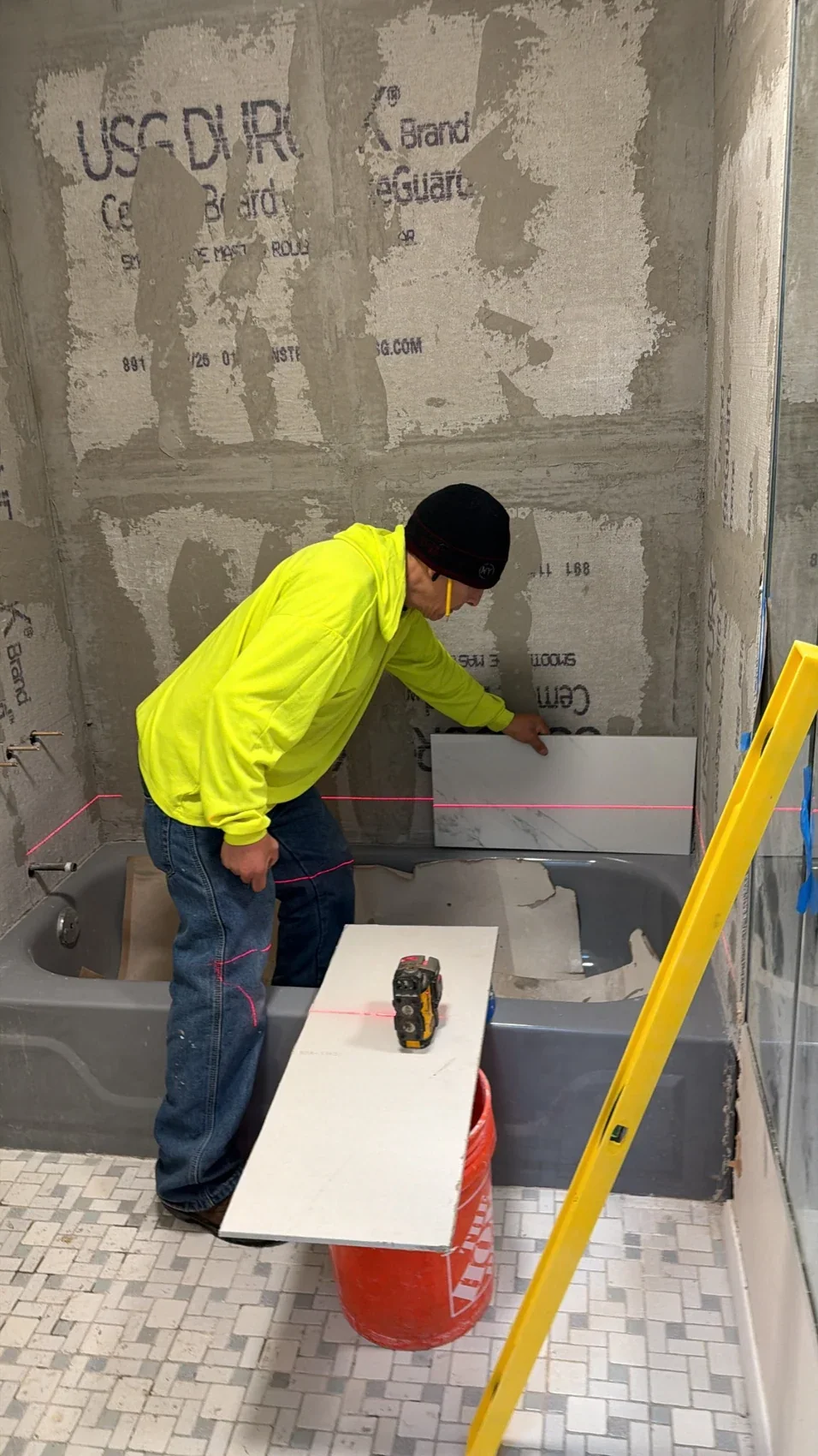Worker positioning a marble-look tile on cement backer board above a bathtub during bathroom renovation